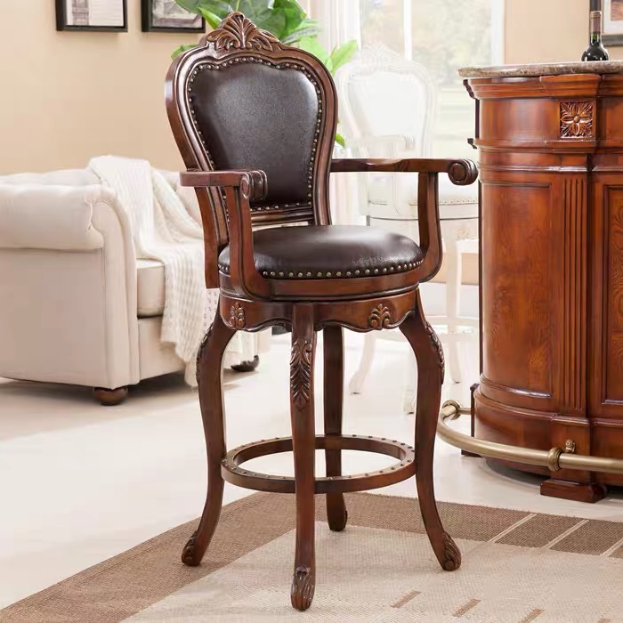 An ornate traditional swivel bar stool from Living Well ZA featuring a rich brown solid wood frame with detailed hand-carved accents on the legs and upper crest. The high-back armchair design includes a matching dark brown genuine cowhide leather seat and backrest trimmed with hand-hammered metallic rivets, positioned on a patterned rug next to a luxury wooden bar counter.