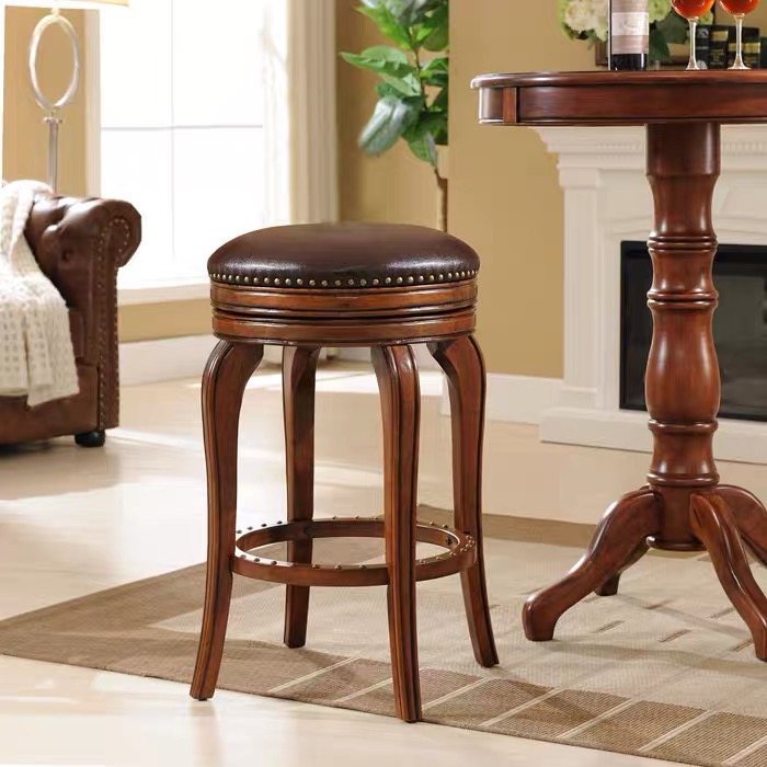 Antique-style backless counter stool with a dark brown solid wood frame, curved legs, and a round coffee-colored genuine cowhide leather swivel seat featuring brass nailhead trim, styled next to a traditional wooden pub table in a warm living room setting.